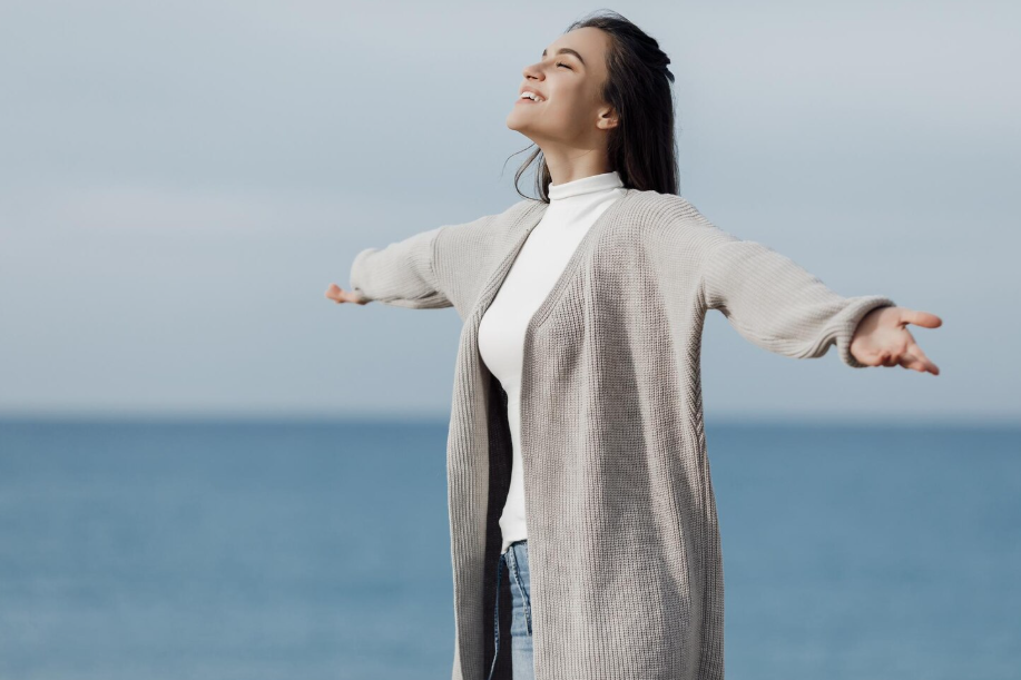 Woman with arms outstretched by the sea, eyes closed, breathing deeply and enjoying a calm moment.