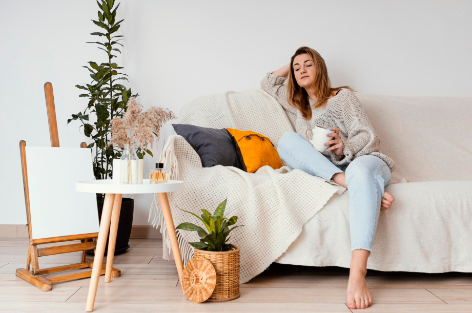 Woman relaxing on a cozy sofa with a soft knit blanket and houseplants in a calm, minimalist living room.