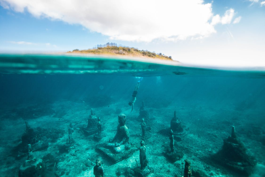 Snorkeler descending toward underwater stone Buddha statues and stupas, illustrating underwater archaeology.