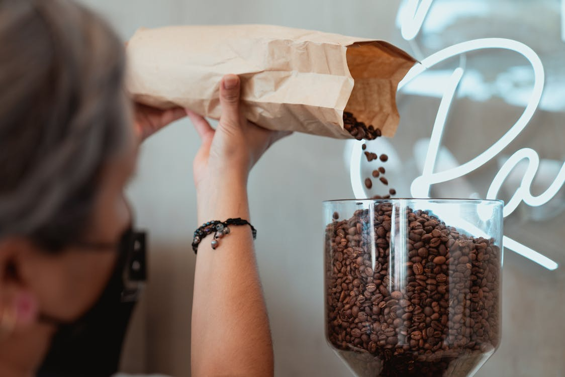 Barista pours roasted coffee beans from a paper bag into a grinder hopper at a café.
