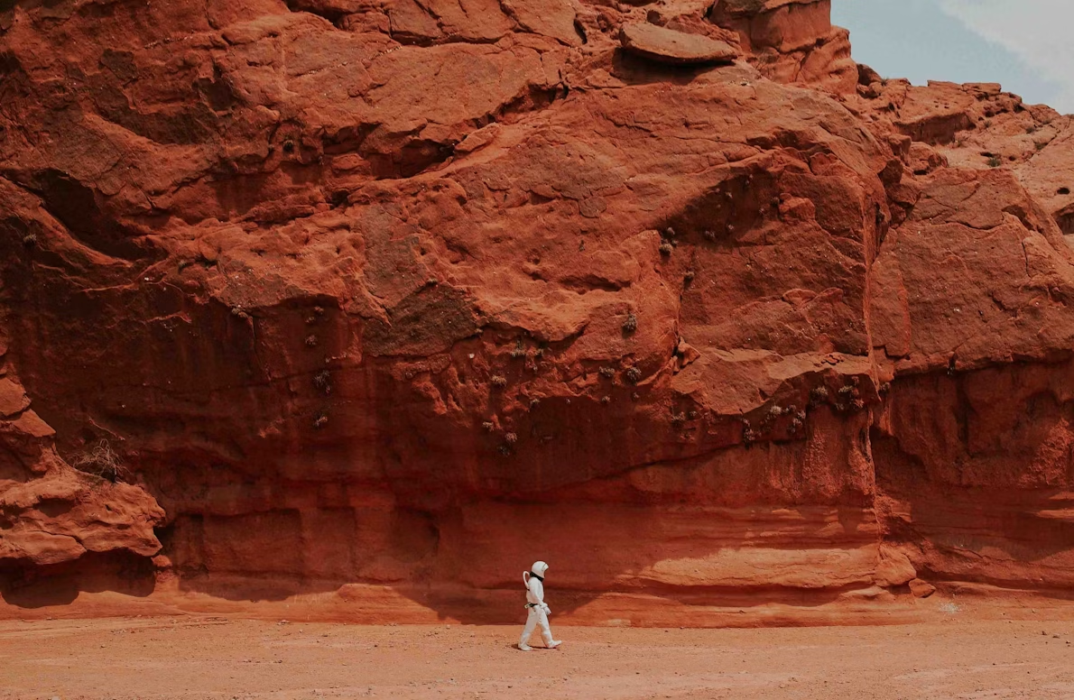 Astronaut in white suit walking beside red sandstone cliff, evoking Mars exploration and survival.